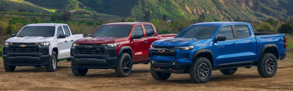 Three 2025 Chevy Colorado trucks — one white, one red, and one blue — parked on red dirt terrain with green mountainous scenery in the background near JK Chevrolet in Nederland, Texas.