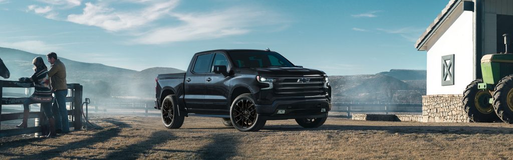 A black 2025 Chevrolet Silverado 1500 with black rims parked beside a farm shed, with mountain views in the background. To the left, a man, woman, and horse stand near a fence; to the right, a green tractor is partially visible near the farmhouse. Scene captured near JK Chevrolet in Nederland, Texas.